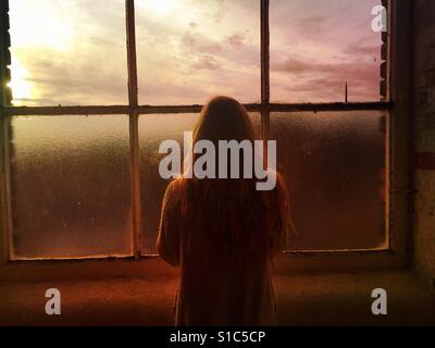 Rückansicht einer einsamen Frau, Blick aus dem Fenster bei Sonnenuntergang Stockfoto