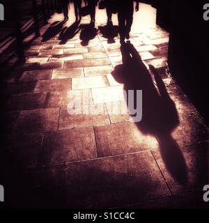 Der lange Schatten von einer Person zu Fuß entlang der Fahrbahn, dicht gefolgt von vier weiteren Personen in der späten Abendsonne. Stockfoto