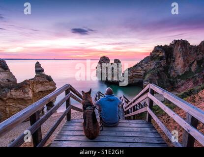 Junger Mann beobachten schönen Sonnenaufgang mit Belowed Hund am Praia de Camilo in portugal Stockfoto