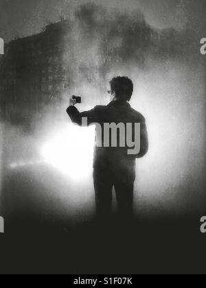 Schwarz / weiß Foto mit dramatische Beleuchtung ein Mann hält seine Kamera-Handy auf Armlänge beim posieren für ein Selbstporträt. Stockfoto