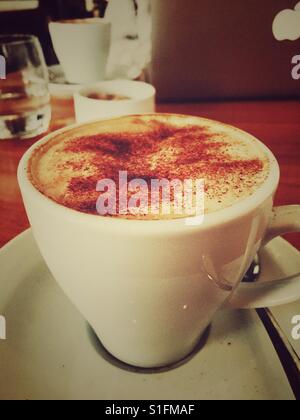 Cappuccino Kaffee auf einer hölzernen Tischplatte in einem Café gegenüber einer Person mit einem MacBook Stockfoto