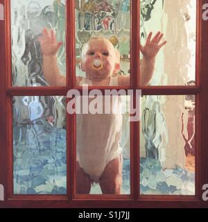 Zehn Monate altes Baby Junge stand gegen eine interne Glastür. Stockfoto