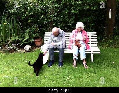 Ein älteres Ehepaar saß auf einer Gartenbank werden von einer freundlichen schwarzen Katze angefahren. Stockfoto