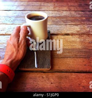 Die Hand eines Mannes mit einem frischen Americano Getränk ruht auf einem Holztisch Stockfoto