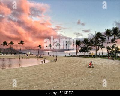 Waikiki Beach und Diamond Head mit rosa Gewitterwolken Overhead bieten schöne Kulisse kleiner Junge Reiten Fahrrad auf Sand von tropischen Palmen gesäumten Lagune, Oahu, Hawaii Stockfoto