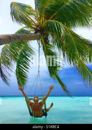Junger Mann genießt den Moment der Freiheit und des Glücks des Urlaubs mit seinen Händen in der Luft hängend Kokospalme in klarem Wasser auf den Malediven im Gange. Stockfoto