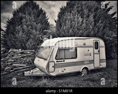 Eine alte, verlassene caravan in der Ecke eines Feldes sitzt, neben einem Haufen von Protokollen. Zwei große, immergrüne Bäume bieten Schutz. Foto - © COLIN HOSKINS. Stockfoto