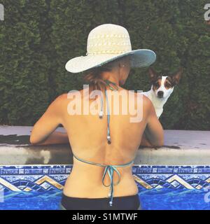 Frau Sonne hat am Rand des Pools mit Blick auf kleinen Hund auf dem Pooldeck. Stockfoto