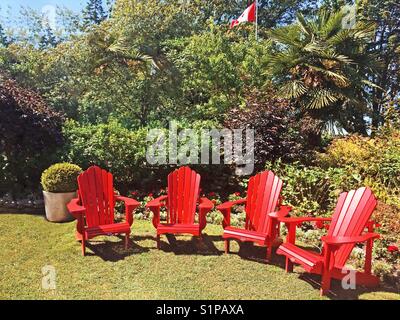 Red Adirondack Stühle leer in einem üppigen Garten, mit einem kanadischen Flagge im Hintergrund. Stockfoto