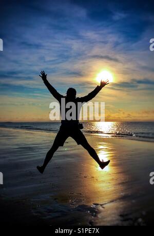 Die Silhouette eines Mannes springen hoch in der Luft auf der sonnenbeschienenen Strand bei Sonnenuntergang mit den Ozean dahinter. Stockfoto