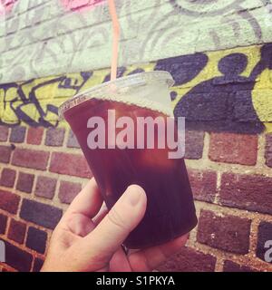 Eine Frau hält ein Glas Eiskaffee vor einer Ziegelmauer mit Graffiti drauf Stockfoto