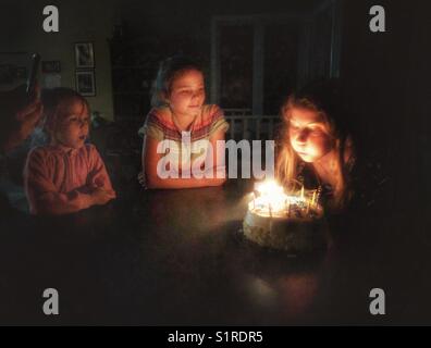 Ein Mädchen, das die Kerzen auf ihrem Geburtstagstorte ausbläst und Schwestern zusieht Stockfoto