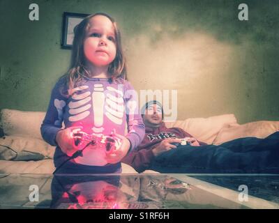 Kleines Mädchen in lila Skelett Schlafanzug holding Controller spielen Videospiele mit ihrem Vater. Stockfoto