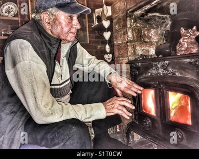 Alter Rentner warm zu halten neben einem Holzofen in seinem Suffolk home. Stockfoto