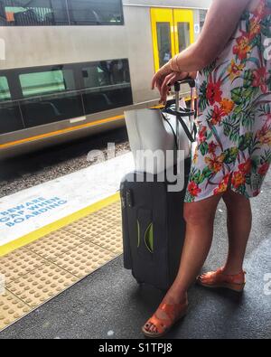 Stilvolle Frau steht wartend am Bahnsteig mit Koffer mit Rädern. Die Reisenden in den Sommerferien mit der Bahn. Dame in hellen Kleid wartet am Bahnhof mit Gepäck. Stockfoto