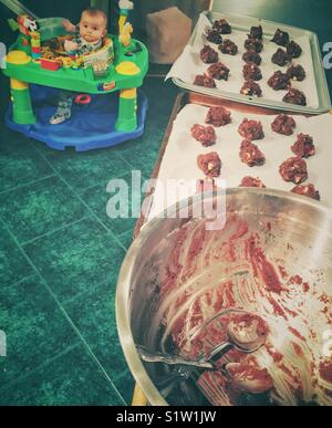 Die Triple Chocolate cookies mit Löffel von rohem Teig auf Cookie Pfannen und Schüssel mit Baby in Activity Center im Hintergrund Stockfoto