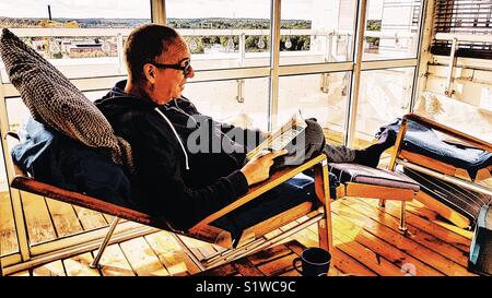 Im mittleren Alter kaukasischer Mann auf Liegestuhl auf der geschlossenen Terrasse ein Buch lesen liegend, Schweden Stockfoto