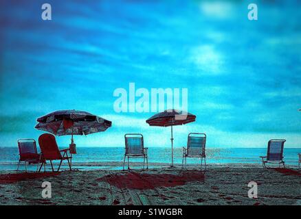 Leere Stühle und Sonnenschirme an einem Sandstrand mit einem ruhigen Meer und Hellblau bewölkter Himmel Stockfoto