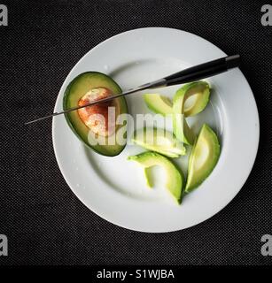 Halbe Avocado mit dem Messer in der Grube und Scheiben auf weiße Platte Stockfoto