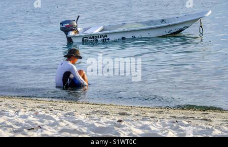 Mann sitzt im Wasser nach Fischen. Sein kleines Boot neben ihm verankert Stockfoto
