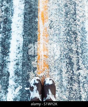 Blick nach unten an der braunen Stiefel der Person, die auf die gelbe Linie in der Mitte der verschneiten Piste Stockfoto