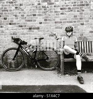 Eine weibliche Radfahrer nimmt eine Pause auf einer Bank mit ihrem Fahrrad in der Nähe Stockfoto