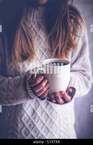 Ein Mädchen in einem gemütlichen, Arran Pullover mit einer heißen Tasse Kaffee oder einem Becher Kaffee in der Hand drinnen. Stockfoto