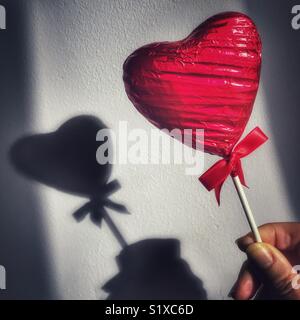 In Herzform, Folie eingewickelt, Schokolade Lutscher, in der Hand einer Frau mit Schatten an der Wand Stockfoto