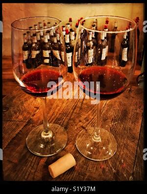 Zwei Gläser mit Rotwein auf einer hölzernen Tischplatte werden durch einen Korken im Vordergrund und viele leere Weinflaschen im Hintergrund in einem Café in Ajijic, Mexiko umgeben. Stockfoto