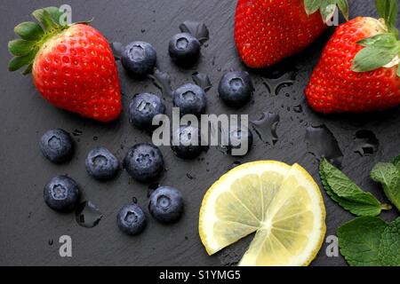 Erdbeeren, Blaubeeren, Zitronenscheiben und Minze mit Wassertropfen auf schiefer Board Stockfoto