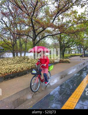 Japanische Frau gekleidet in Rot reiten ihr Fahrrad ihrem Schirm halten an einem regnerischen Tag Stockfoto