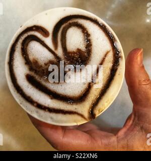 Frau mit einem Schaumigen weißen Kaffee mit einem Smiley Herz in einem Takeaway Schale Stockfoto