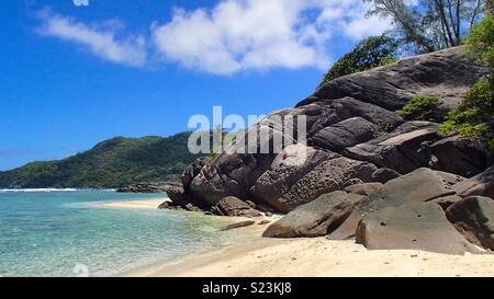 Grau Schwarz Rock teilen Sie die Abschnitte der Strand an der Küste der Seychellen in dieser schönen Badebuchten mit türkisfarbenem Wasser, grünen Palmen, weiße Sandstrände und blauer Himmel Stockfoto