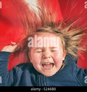 Portrait von Happy toddler girl Gesicht, während sie rutscht eine rote Folie mit geschlossenen Augen, den Mund offen und Haar kleben von statischer Elektrizität Stockfoto