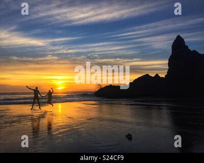 Paar springen halten sich an den Händen, als die Sonne im Hintergrund auf Piha Beach, Auckland, Neuseeland Stockfoto