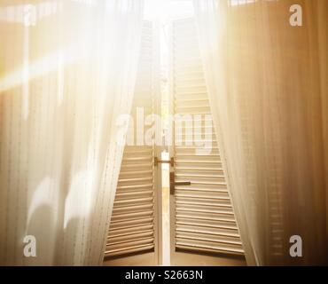 Sonnenlicht gefiltert durch die Fensterläden aus einem offenen Fenster mit Vorhängen aus weißer Baumwolle Stockfoto