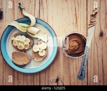 Roggen Toast mit Erdnussbutter und Bananenscheiben auf einem Teller mit einer geschälten Banane neben einem Glas Erdnussbutter mit Messer Stockfoto