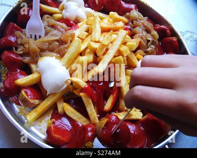 Eine traditionelle Berliner Teller Pommes mit Currywurst und gebratene Zwiebeln mit der Hand eines Kindes erreichen für die Bundesrepublik Jugoslawien Stockfoto