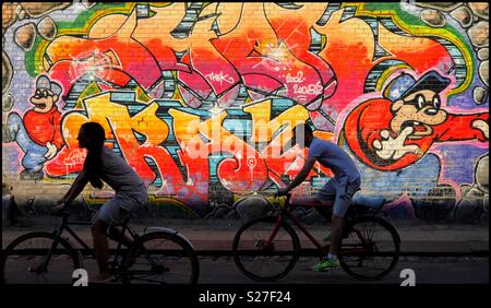 2 Radfahrer fahren an einem bunten Stück von Street Art auf der Seite eines großen Wand gemalt. Es ist Sommer irgendwo in Europa. Foto - © COLIN HOSKINS. Stockfoto