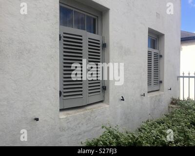 Vorderansicht des Hellgrau Backsteinhaus mit zwei Fenstern die Hälfte von Malte mittelgrau Fensterläden aus Holz Straße im exklusiven Vorort und grünen Pflanzen abgedeckt Stockfoto