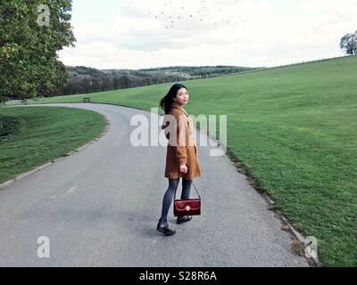 Ein glückliches und modische Chinesischen oder asiatischen Frau in einem Camel Mantel und eine rote vintage Handtasche Blick zurück beim Gehen in Bügel Newsam, Leeds, West Yorkshire, England, Großbritannien Stockfoto