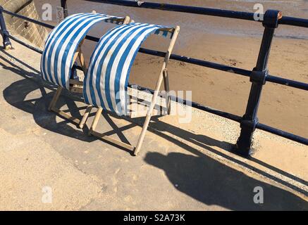 Zwei blau-weiß gestreifte Liegestühle auf einer windigen Promenade Stockfoto