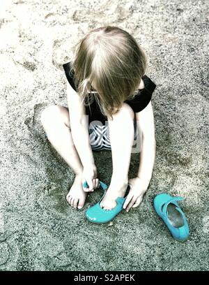 2 Jahre alten Mädchen sitzen im Sand ihre Schuhe setzen auf Stockfoto