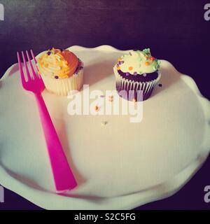 Bunte Vanille und Schoko Cupcakes mit Zuckerguss und Streuseln auf eine weiße Platte mit einem rosa Gabel serviert. Stockfoto