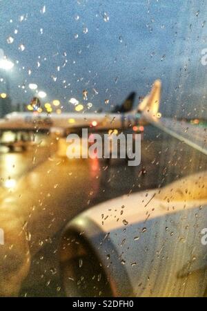 Regentropfen auf dem Flugzeug Fenster beim Warten auf Start- und Landebahn bei Nacht, Flugzeug im Hintergrund. Manchester, Großbritannien Stockfoto