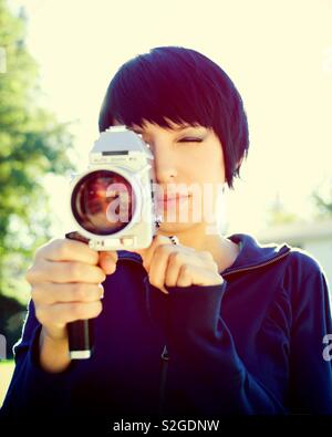 Nettes Mädchen mit kurzen Haaren in ihren frühen Zwanzigern Holding ein Vintage Super 8mm Film Kamera im Freien an einem sonnigen Tag. Foto hat eine Farbe kreuzte Prozess Wirkung. Stockfoto