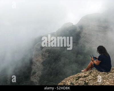 Das Mädchen sitzt auf einem Berg mit dem Nebel eines gefallenen Wolke und steuert die Drohne. Stockfoto