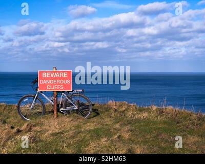 Fahrrad gebunden an eine Warnung Wegweiser auf den Rand einer hohen Klippe mit Blick auf das Meer an einem sonnigen Tag. Stockfoto