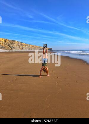 Junge Frau macht einen Handstand auf einem großen Sandstrand, South Wales, April. Stockfoto