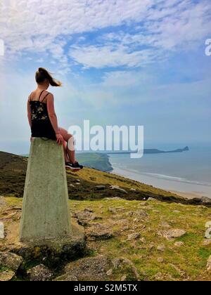 Junge Frau sitzt auf der "Trig Point von rhossili Downs genießen die Aussicht oben Rhossili Bay, Gower, Swansea, Wales. Stockfoto